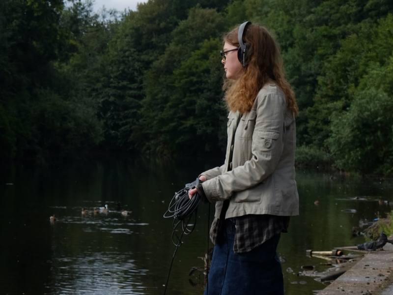 A woman stands by a river wearing and holding equipment which helps to capture sound.