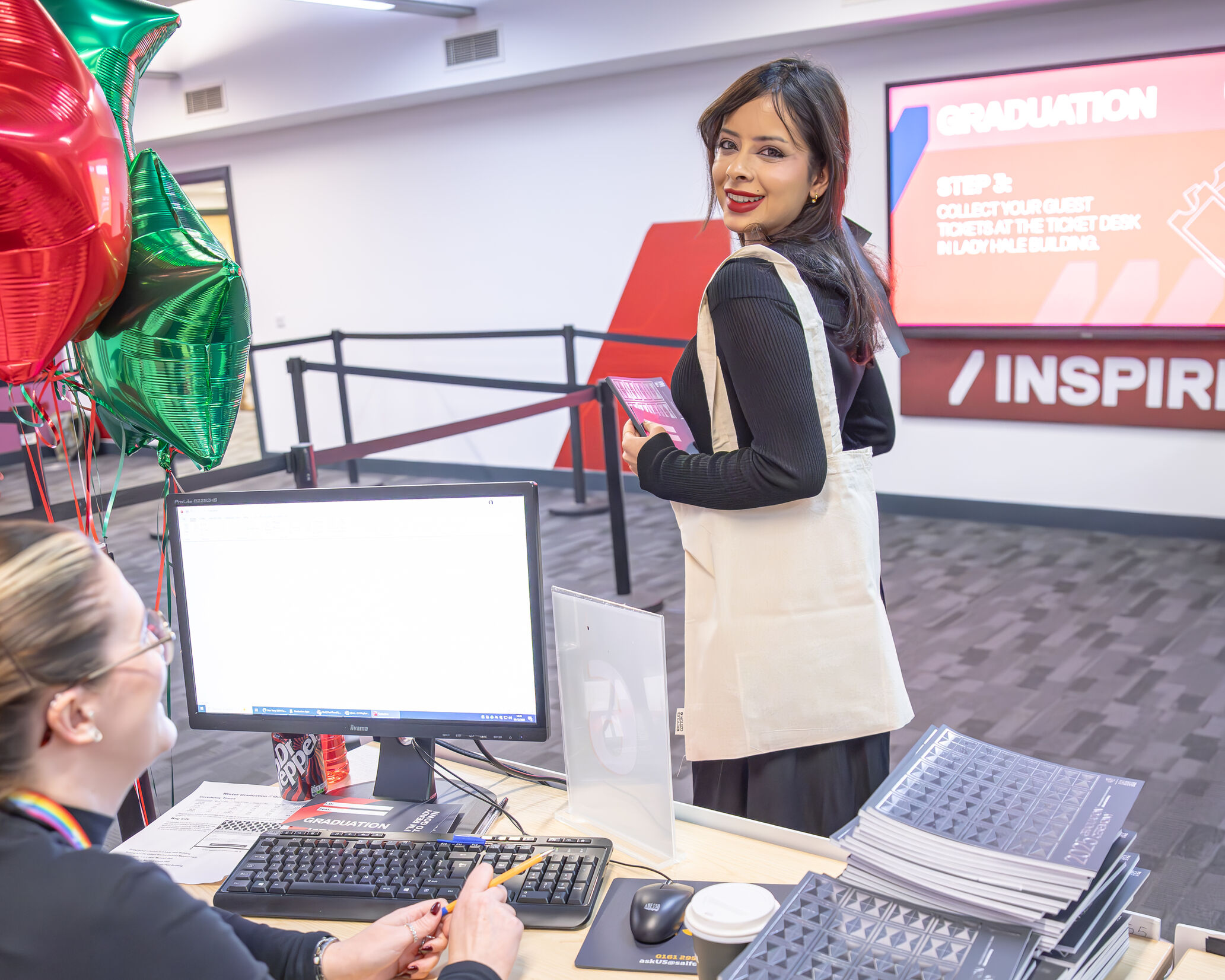 Person checks in at a graduation event desk decorated with red and green balloons, with graduation signage visible in the background