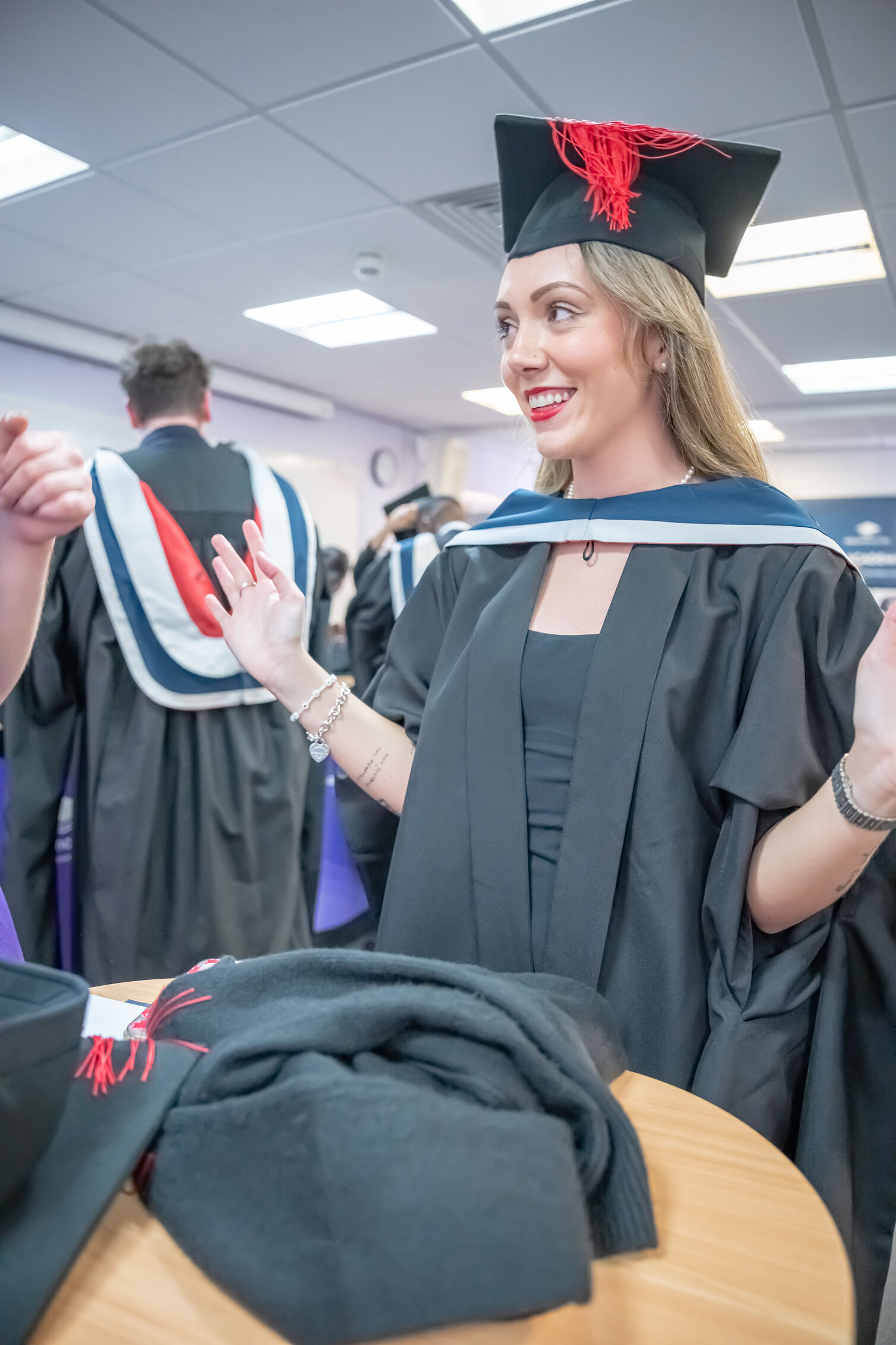 Person wearing a graduation gown and mortarboard with a red tassel stands beside a table with folded academic gowns, interacting with others at an indoor graduation ceremony.