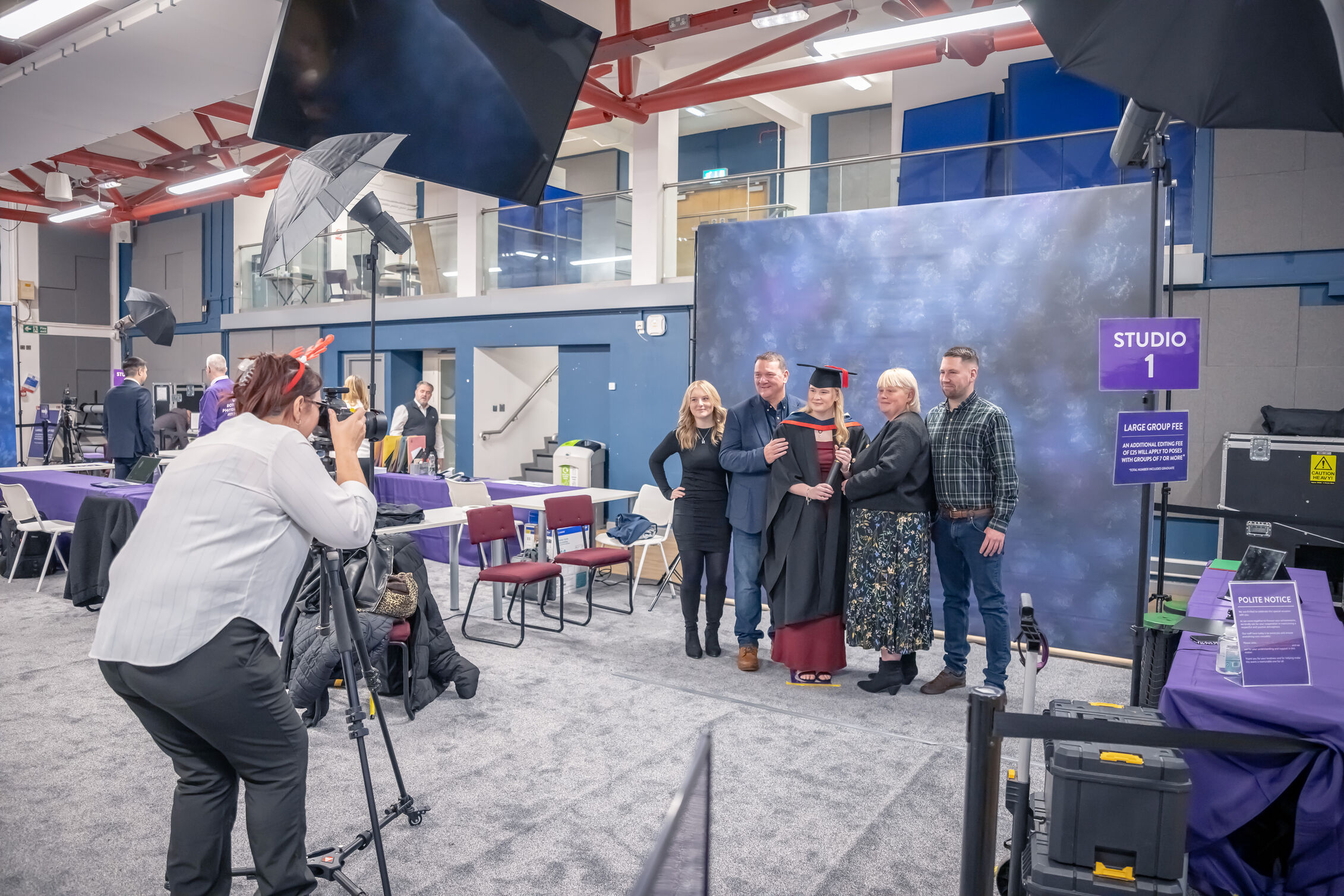 Photographer taking graduation portrait photos of a graduate and family members in a studio setup with lighting equipment and a backdrop at a graduation venue.