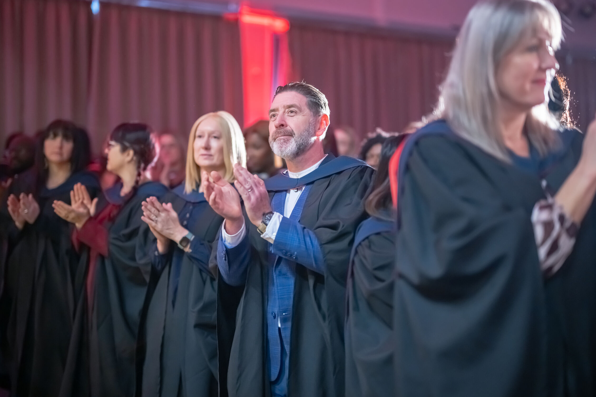 Graduates in graduation gowns standing and applauding during their ceremony.