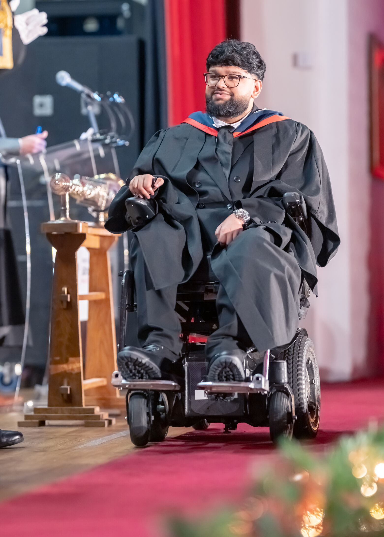 Person wearing graduation robes seated in a powered wheelchair during a graduation ceremony, positioned on a stage beside a lectern with microphones. Red curtains and a red carpet are visible in the background.
