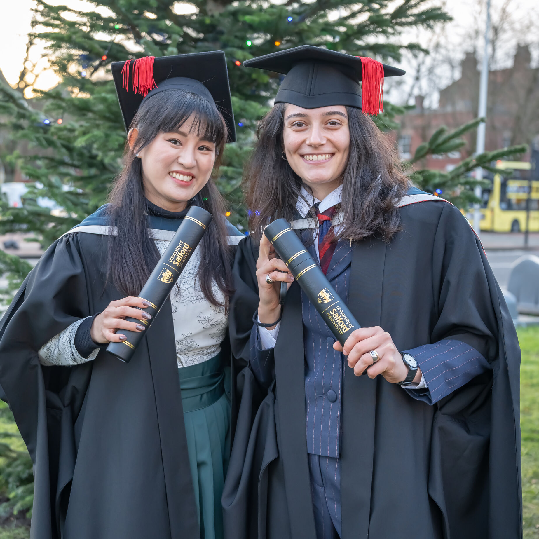 Person in a red graduation gown and academic cap holds a bouquet while posing for a selfie with another person in a park.
