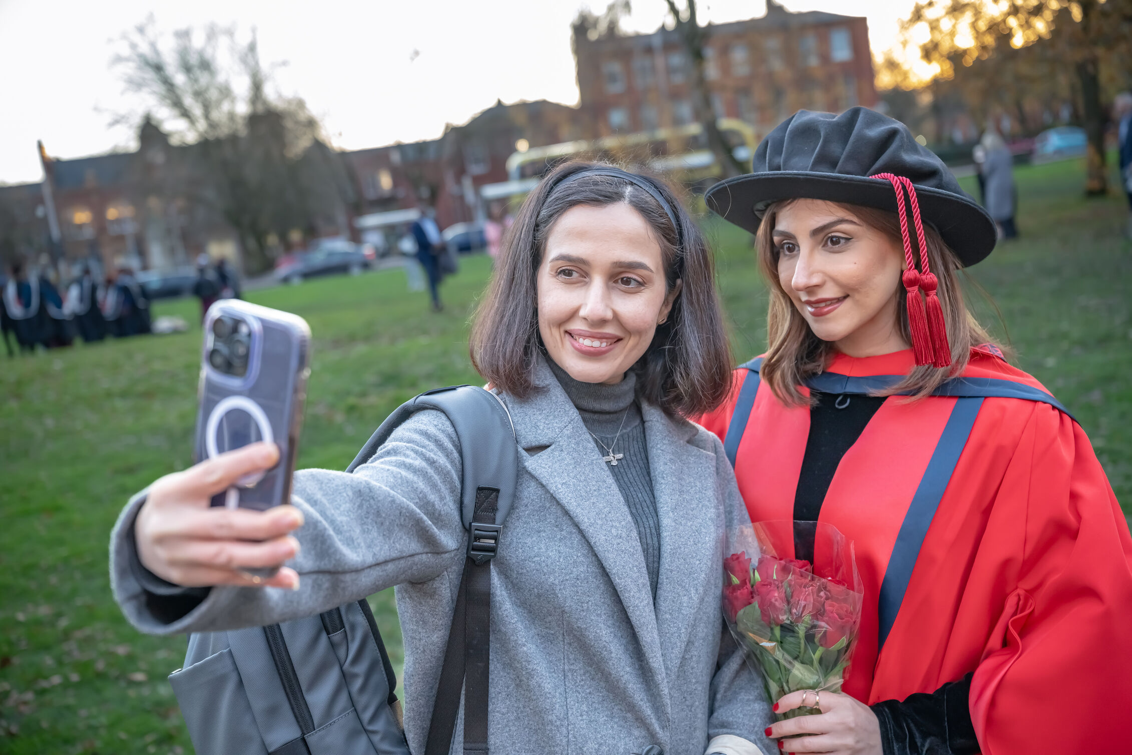 Person takes a selfie with a graduate in academic robes holding a bouquet of roses outdoors.