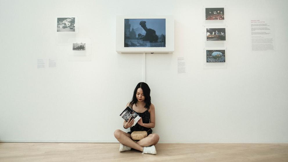 A girl sits on the floor in front of a white wall which contains a digital screen and several photographs