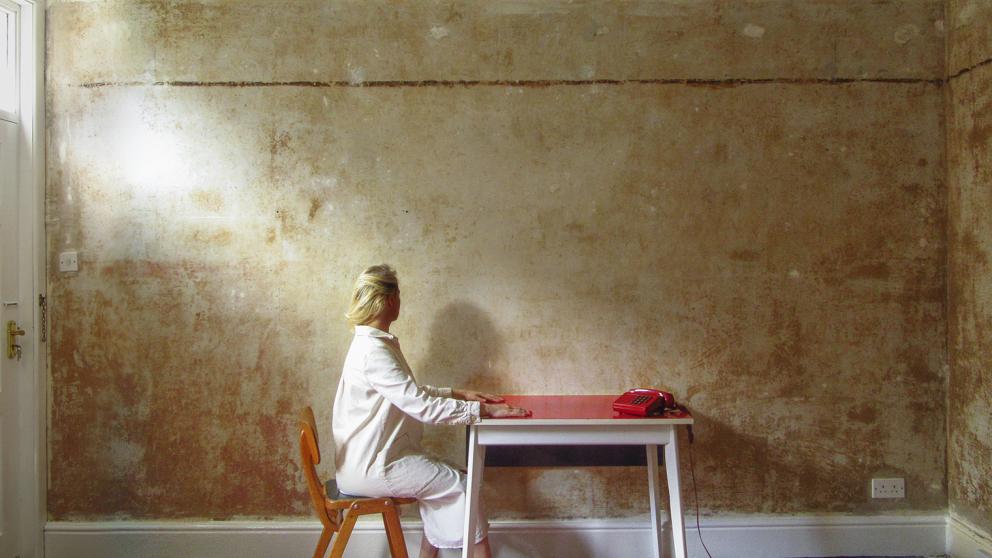 Graduate Lasma Poisa sitting at a table with a red telephone against a rustic wall