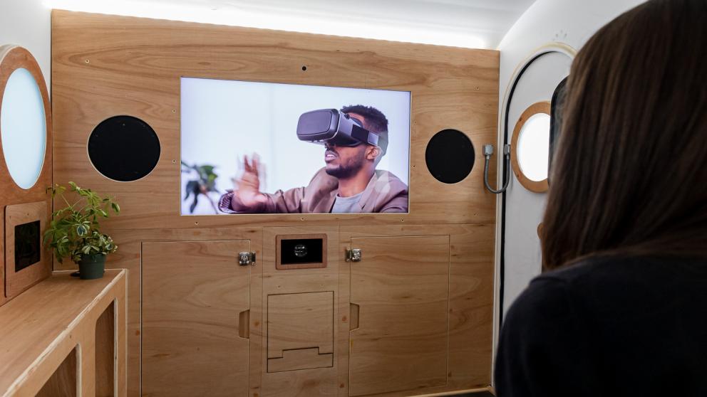 Image shows the interior of the caravan from the future with a big screen and a person watching