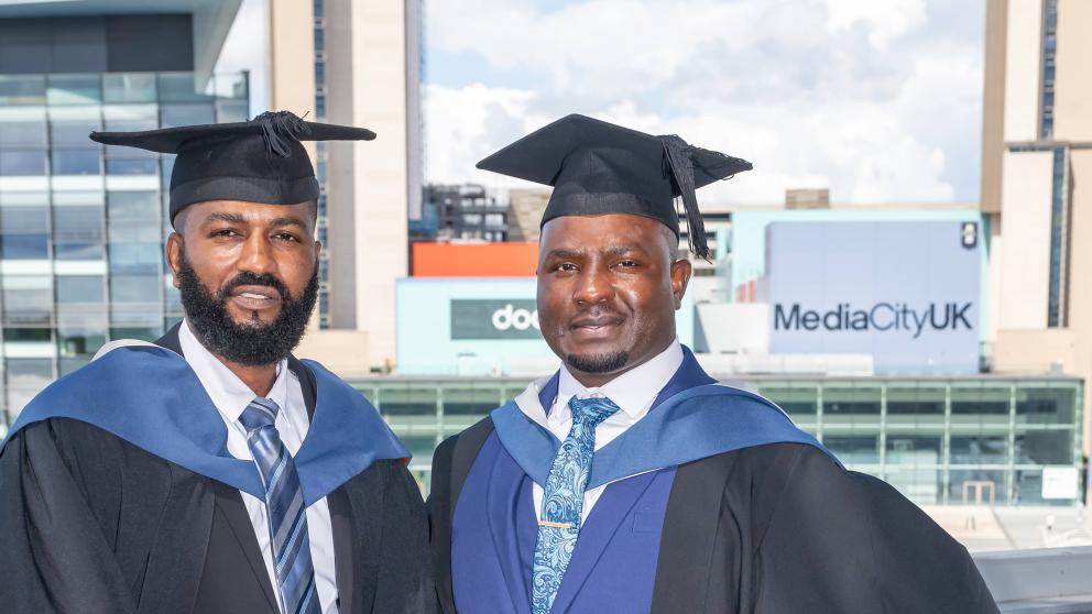 Two men in suits and graduation attire posing for photograph. Big billboards behind them read 'Dock 10' and 'Media City UK' 
