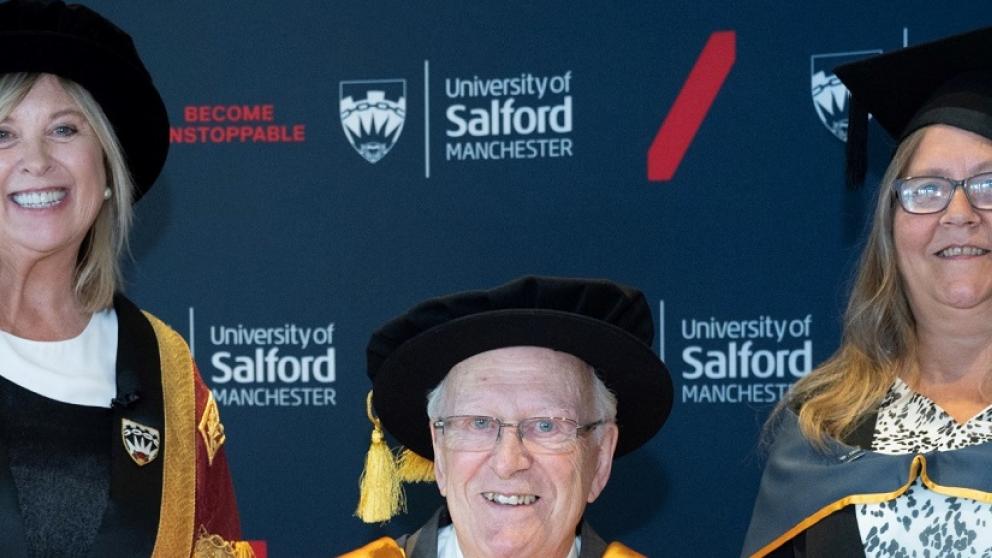 Two women stand next to a man, all in graduation attire in front of wall with University of Salford logo 