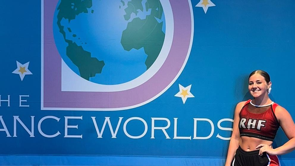 A cheerleader posing in front of a blue with banner containing text: 'The Dance Worlds'.