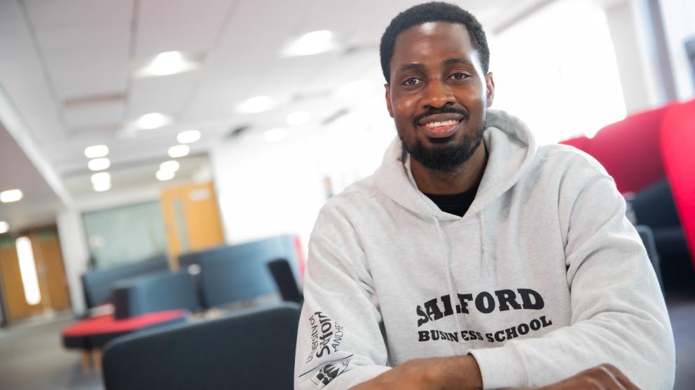Man in grey University of Salford hoodie sitting in a room with several different seats. 