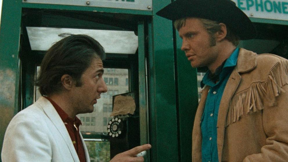 Shot from Midnight Cowboy movie, man in white suit faces man dressed as a cowboy in front of a phone box. 