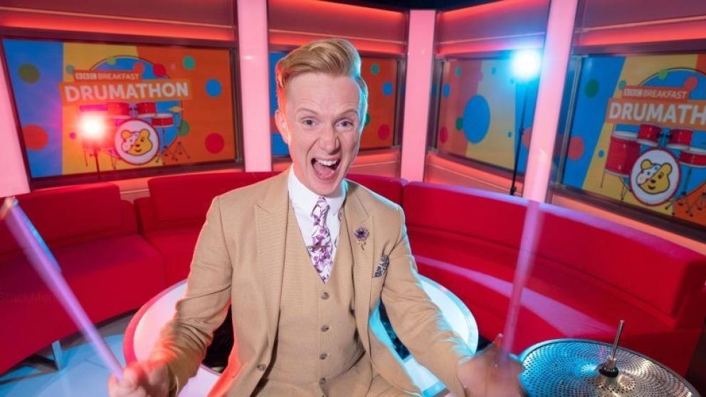 BBC's Owain Wyn Evans posing at a drum kit in BBC TV studios. Screens in the background read: 'BBC Breakfast Drumathon' 