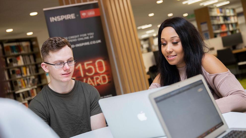 A couple of students looking at the screen of a MacBook together, inside the university’s Clifford Whitworth Library