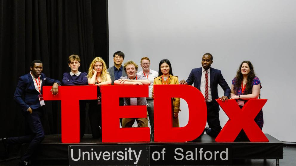 TEDxUniversityOfSalford sign with speakers behind it.