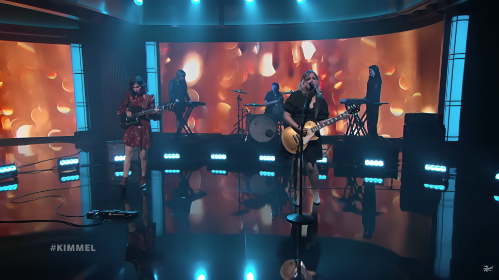 A band of five musicians perform on a stage lit up orange