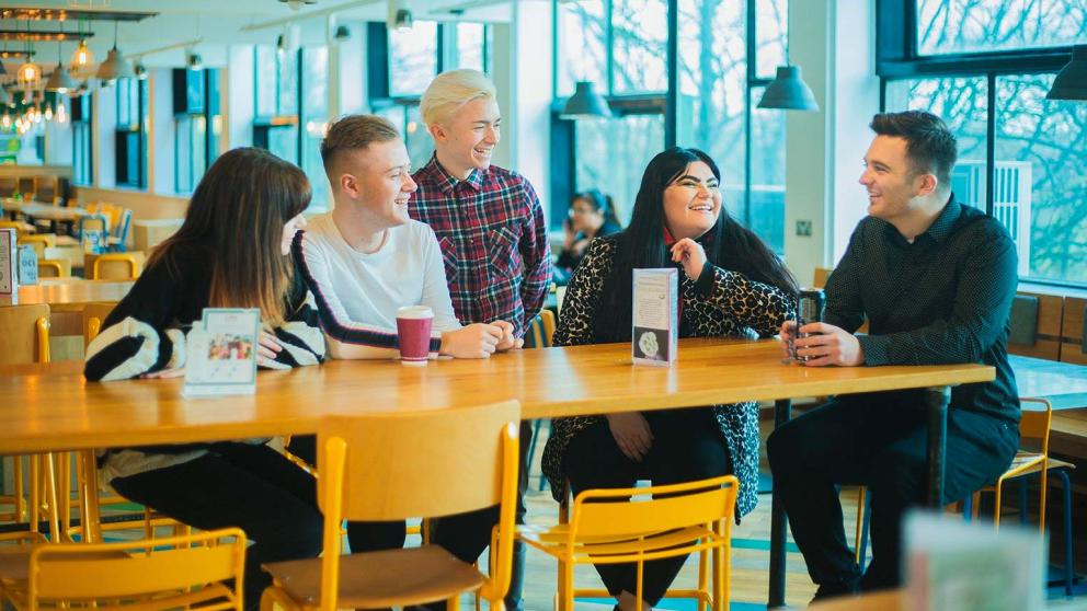 Five students socialising around a table in Maxwell Café, University of Salford