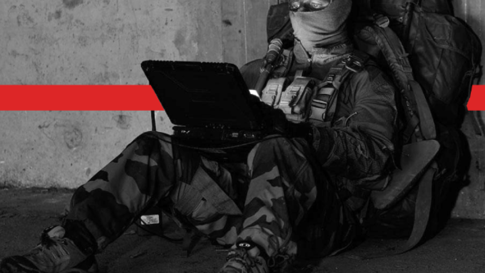 Man in forces uniform sat on ground using electronic device, wearing backpack and face covered.
