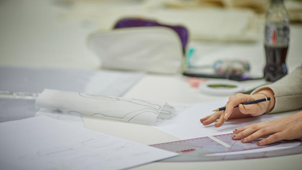 A student’s hands sketching on a desk, with designs on paper laid out around her
