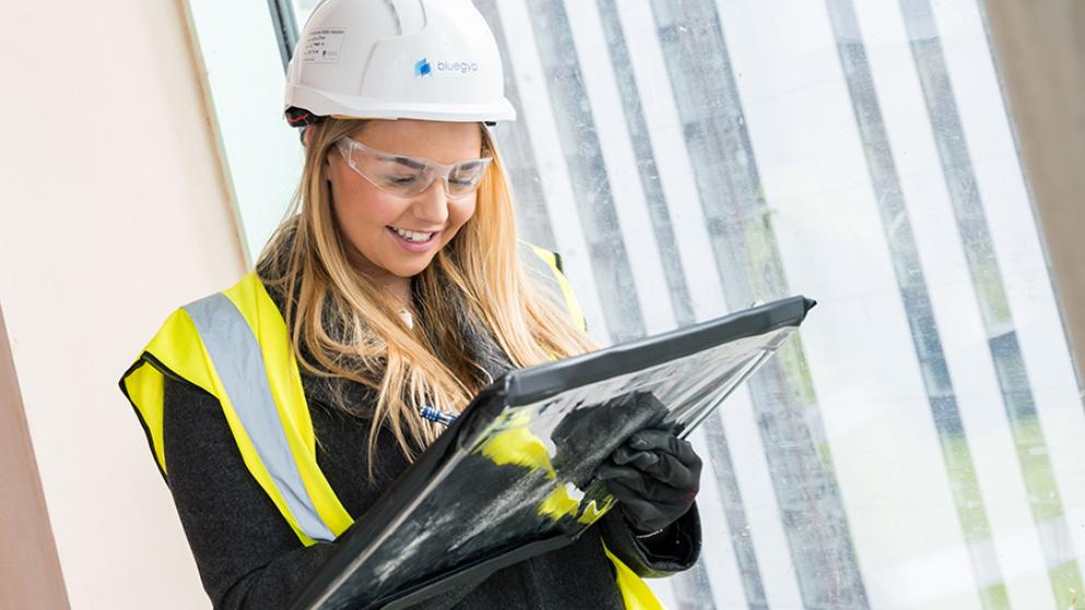 A student in a hard hat, safety goggles, and high-vis jacket smiling and writing on a large pad