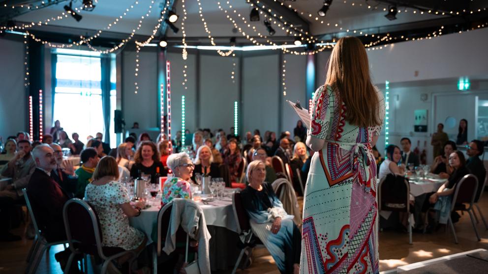 A woman addresses a seated crowd from a stage