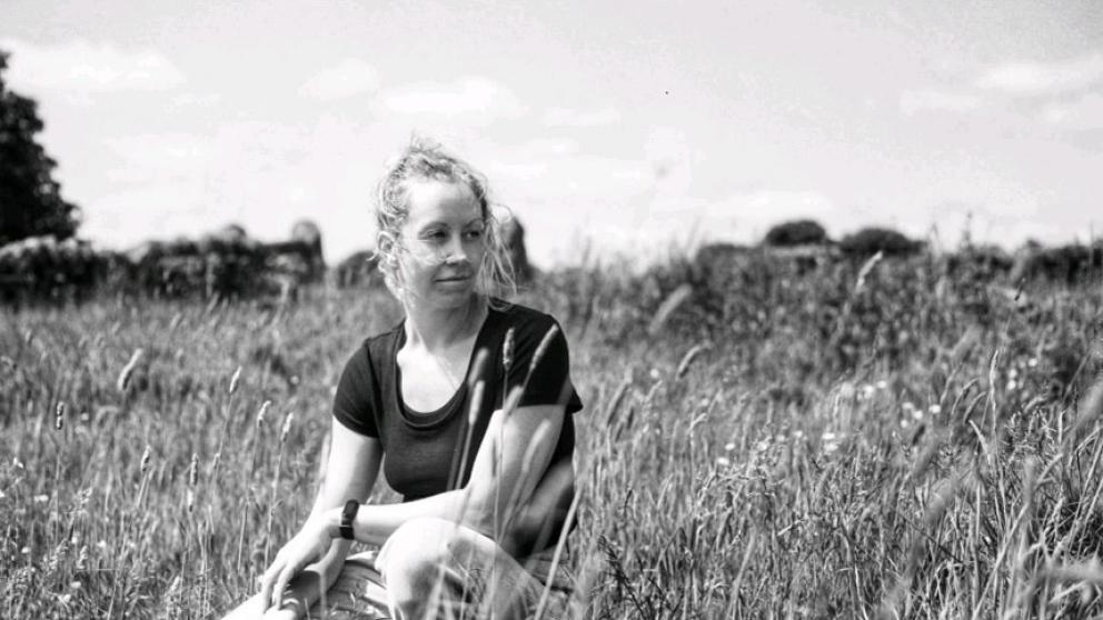 Black and white photo of Dr Emilie Whitaker outdoors amongst some large blades of grass.