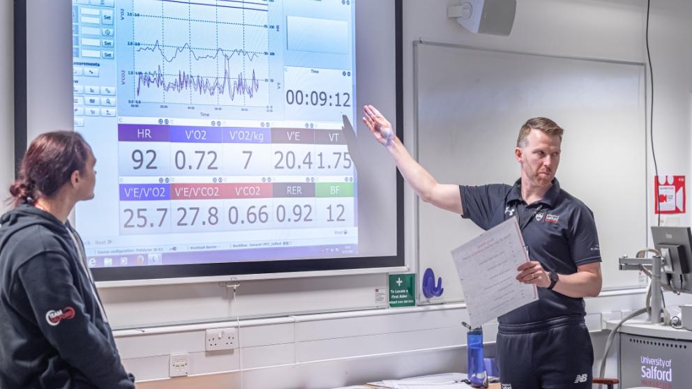 A lecturer, dressed in black, standing in a classroom pointing at a large screen displaying data and graphs related to cardiopulmonary exercise testing (CPET).