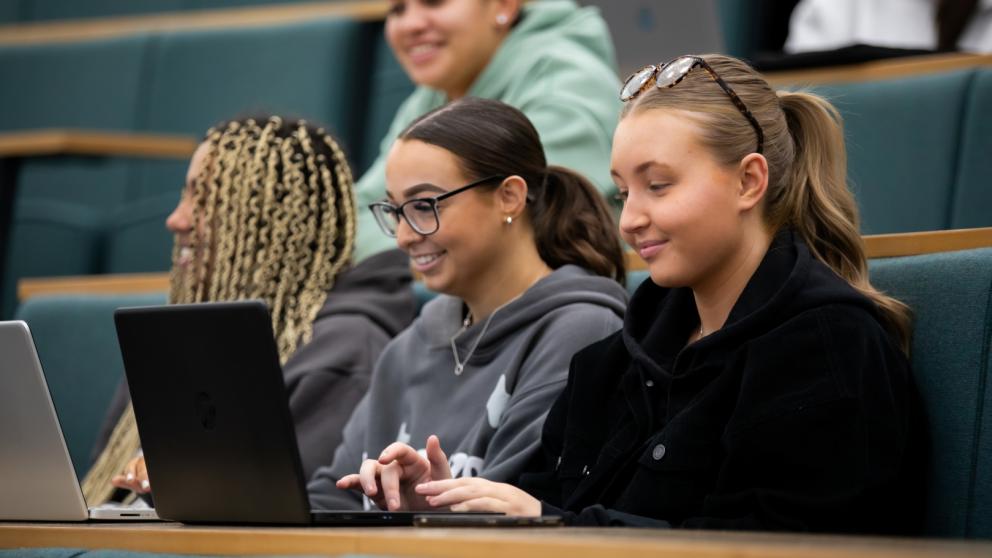 Three students working on laptops in a tiered lecture theatre