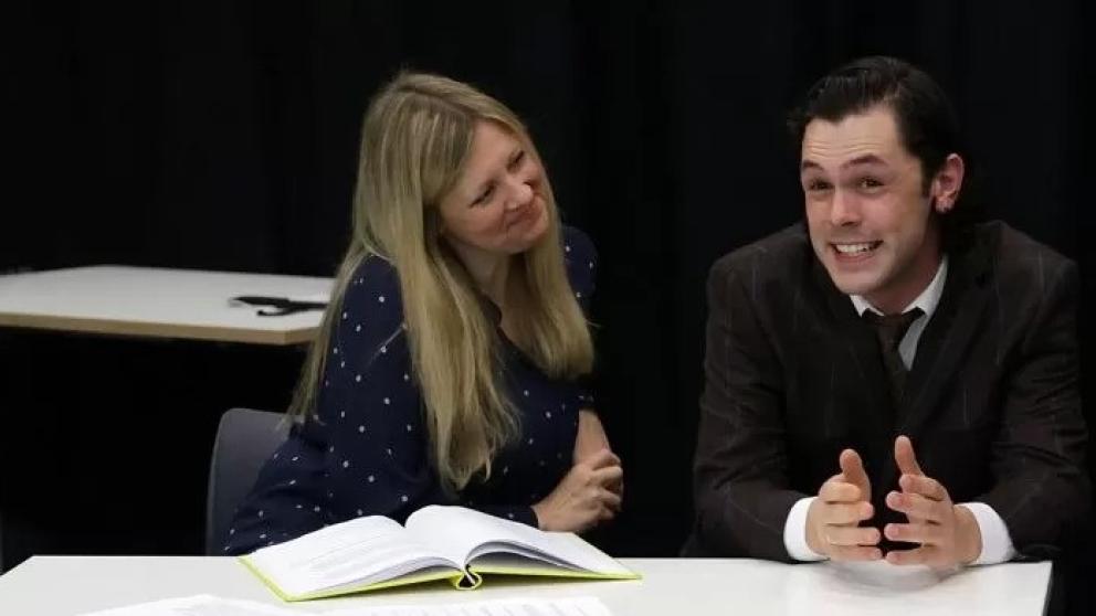 Two performers face an audience sat at a table on a stage 