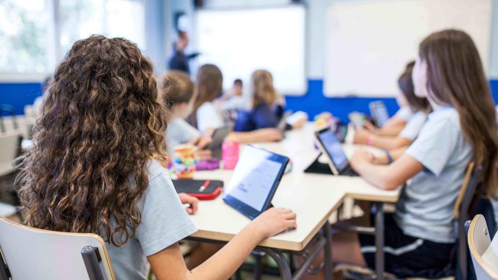 Children are shown using ipads in a classroom environment. They are facing away from camera towards the teacher at the front of the room.