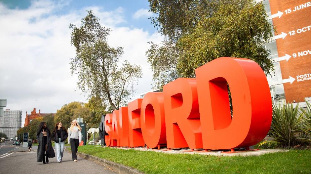 Three female students walking down the street next to a sign which says Salford in big red letters