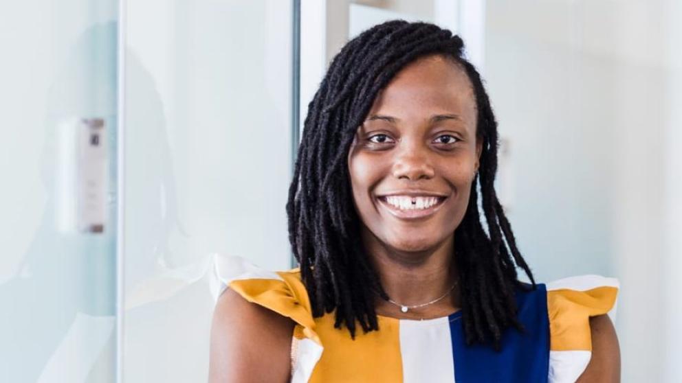 A headshot of Deborah wearing a yellow, blue and white dress