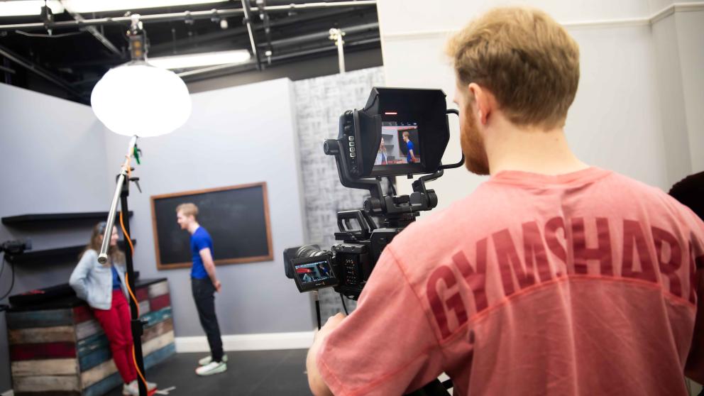 A person behind the camera filming two actors in the TV studio