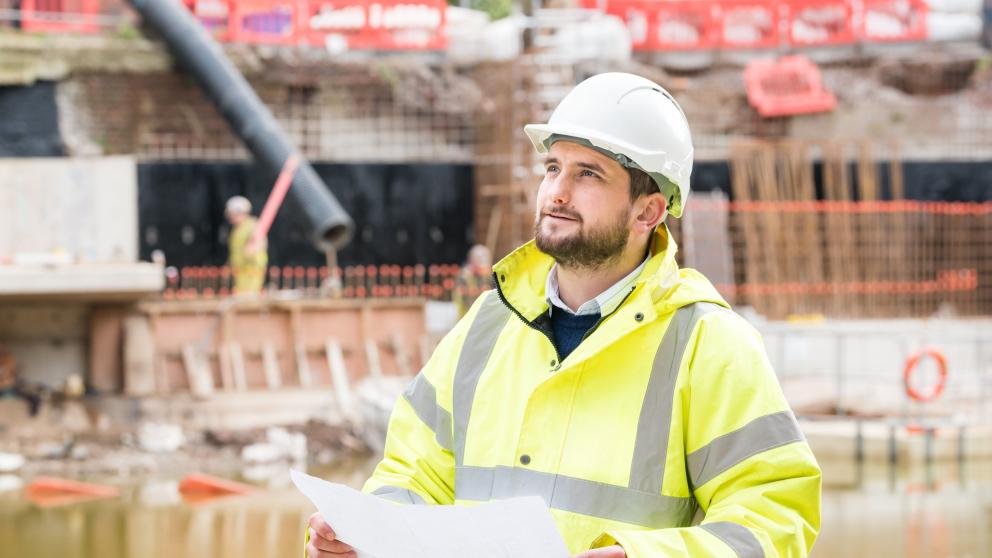 Male construction apprentice on site in safety gear inspecting plans