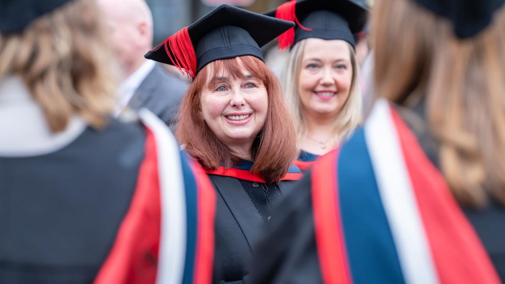 Graduate in Graduation cap and gown smiles at the camera amongst a group of other Graduation 