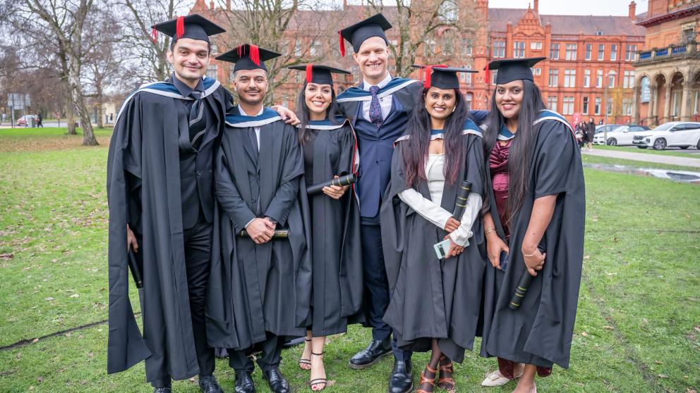 Group of graduates in graduation hats and gowns stand together and smile outside the Peel Building 