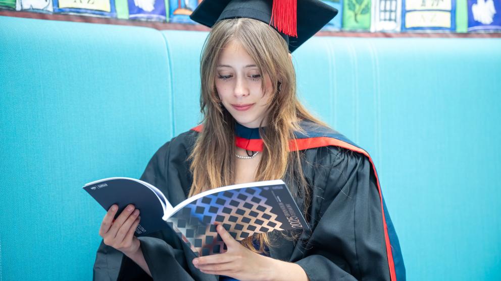 Female Graduate in Graduation cap and gown sits and reads the Graduation Brochure which is navy blue with silver foil diamond patterns
