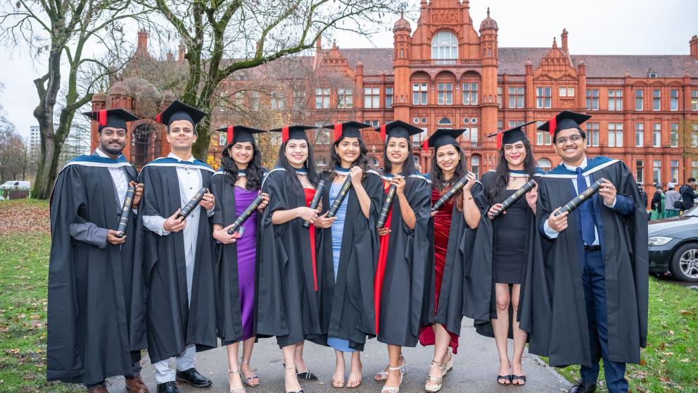 A group of nine graduates in caps and gowns each hold a black scroll with a Gold University of Salford logo on. They stand outside Peel Hall and smile