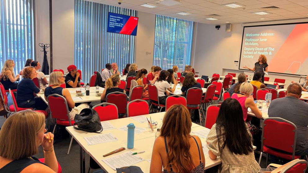 A presenter stands at the front of a busy room of event attendees. The presenter, Professor Jane McAdam, smiles as she addresses the room.