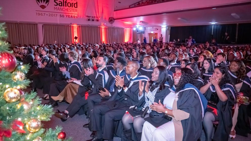 Picture of students in graduation gowns clapping during a ceremony. A Christmas tree can be seen in the corner of the frame.