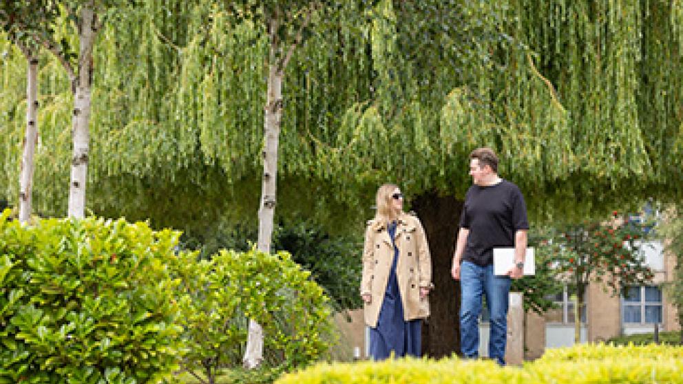 wo colleagues walk outdoors at the University of Salford surrounded by lush greenery and weeping willows. One wears a beige trench coat and sunglasses; the other, in a black shirt and jeans, holds a laptop. They appear to be in discussion