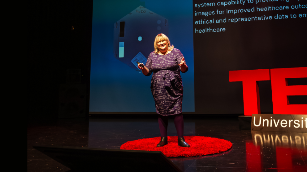 Dr Katy Szczepura presenting a talk on stage. She is smiling and holding her hands wide as she speaks. A banner behind her reads: "TedX University of Salford".