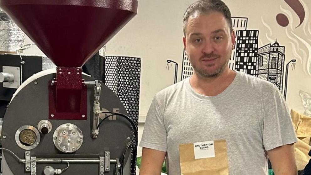 Three people standing in front of a coffee bean grinding machine with one holding a bag of coffee beans