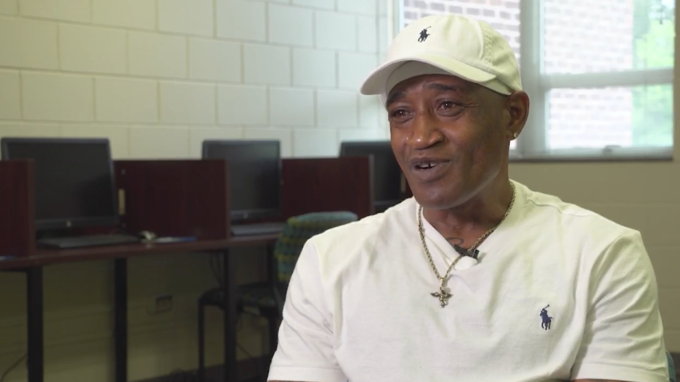 A man in a white shirt and white baseball cap speaks to a camera