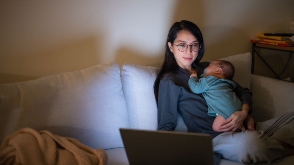 A mum sat working on her laptop while holding her child who is sleeping