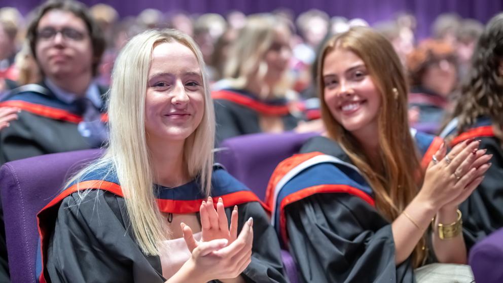 Two graduated is black gowns with red, grey and dark blue hoods smile and clap in the audience of a ceremony.