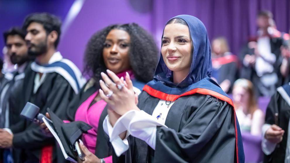 A graduate stands during their ceremony and applauds while looking ta the stage smiling.