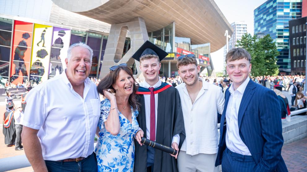Graduate in a Graduation gown smiles in front of the Lowry theatre. Four guests stand with the graduate smiling and laughing.