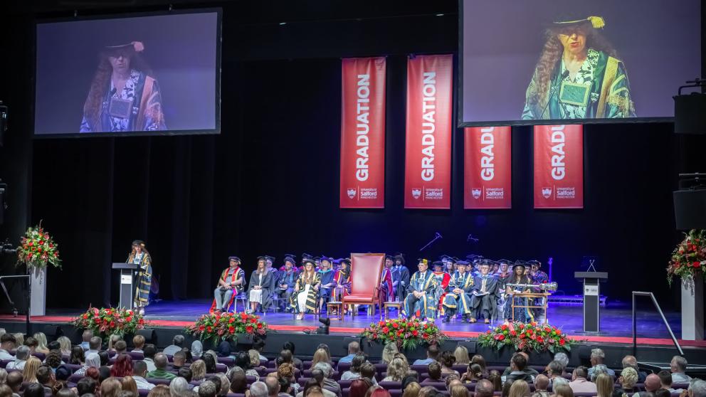A crown can be seen watching a Graduation ceremony. A procession sits on stage in various gowns. Red banners reading graduation can been seen hanging in the back of the stage. 