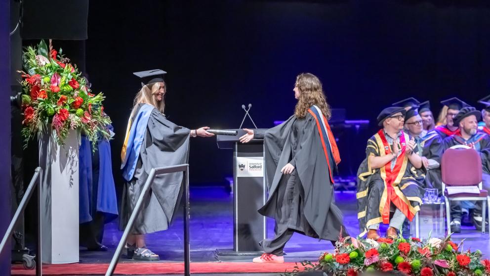 A graduate in a black gown with a red, grey and dark blue hood, crosses the stage at their Graduation. The Graduate holds out their hand to receive a black scroll with gold detailing on it. The scroll is handed to them by a member of staff wearing a black gown with a yellow and blue hood and mortar board hat. A procession sits on stage behind the pair and applauds. A large bouquet of flowers can be seen at the end of the stage on a white pillar.